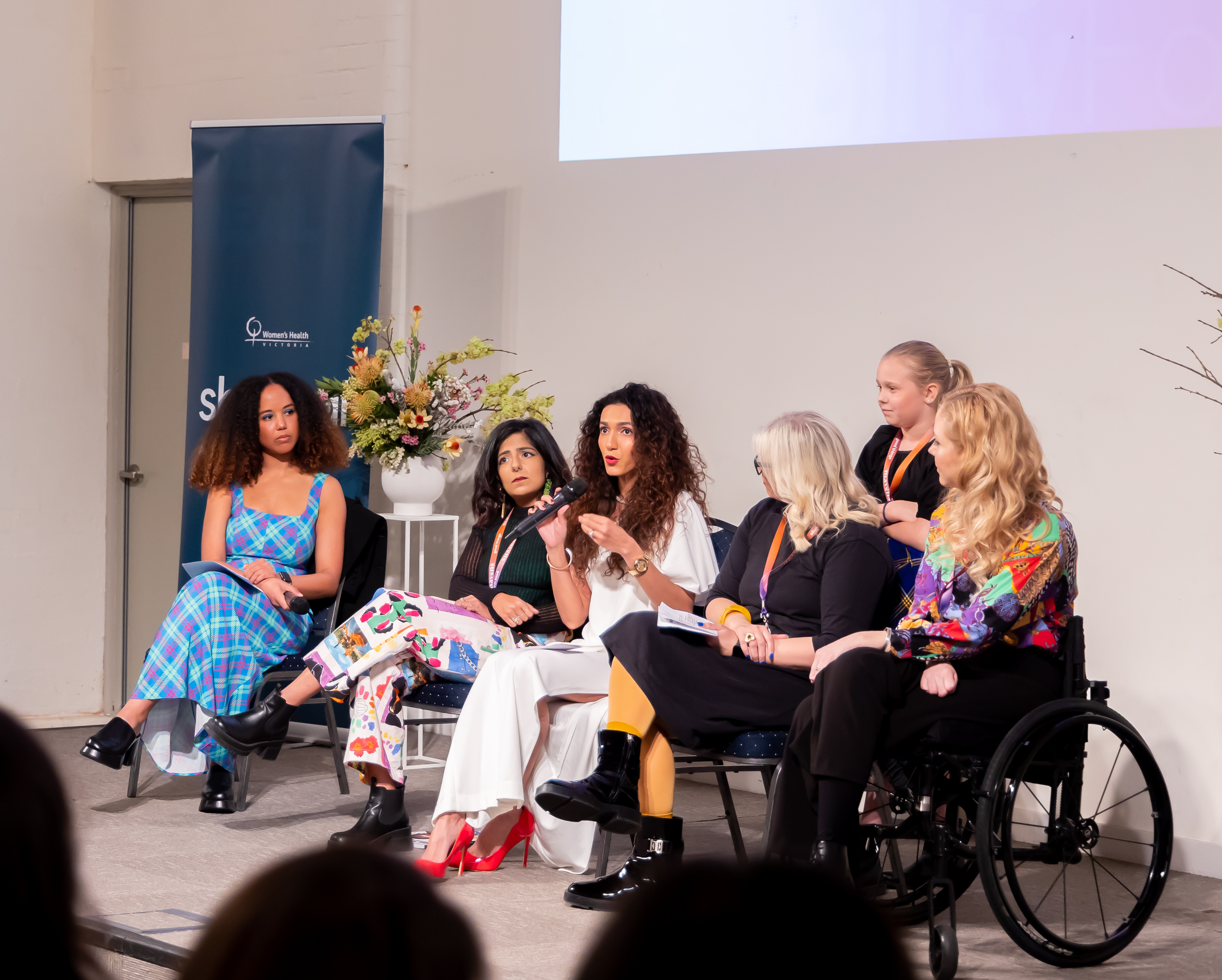 Six women sit on a panel discussing inclusion, with one speaker holding a microphone while others listen attentively, including a panelist in a wheelchair.